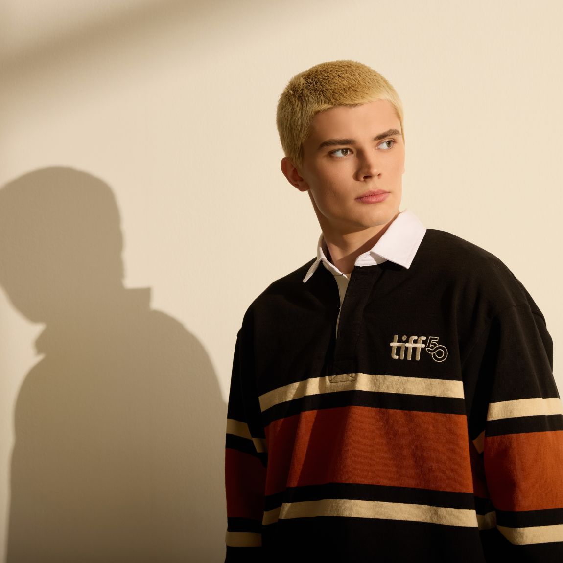 Person standing against beige background wearing orange and black striped rugby sweater with white TIFF 50 logo embroidery at chest.