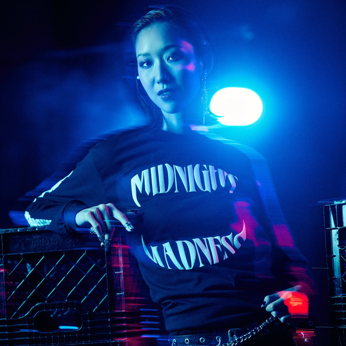 Person with hand at back of neck wearing a red MIDNIGHT MADNESS branded t-shirt and various silver necklaces, surrounded by blue and red hues.