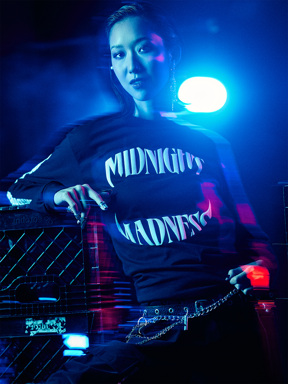 Person with hand at back of neck wearing a red MIDNIGHT MADNESS branded t-shirt and various silver necklaces, surrounded by blue and red hues.