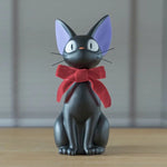 A black cat figurine representing Jiji from Kiki's Delivery Service sits upright with large eyes, purple inner ears, and a prominent red bow tied around its neck.
