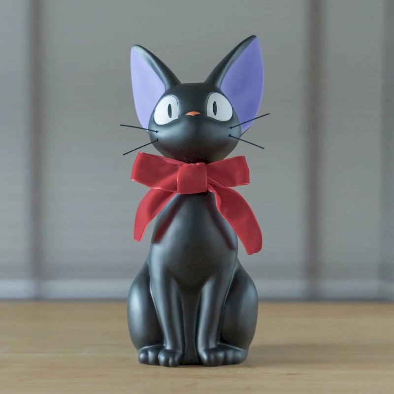 A black cat figurine representing Jiji from Kiki's Delivery Service sits upright with large eyes, purple inner ears, and a prominent red bow tied around its neck.
