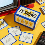 A box for the party game "TV Shows Poorly Explained" sits on a yellow surface surrounded by game cards featuring humorous, oversimplified synopses of popular television series.