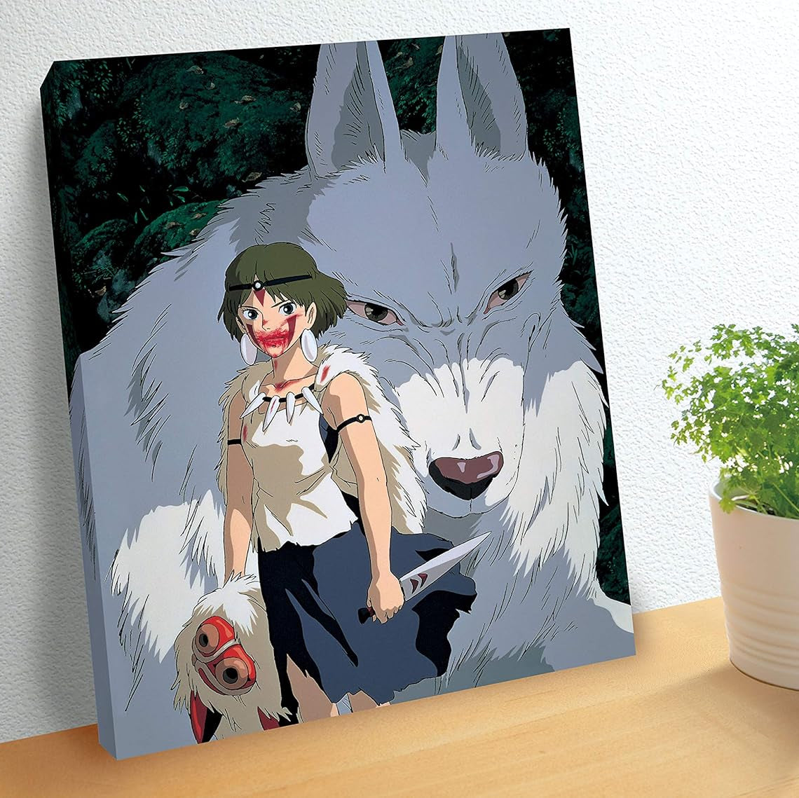 A framed art print of Princess Mononoke standing with Moro the wolf god leans on a wall beside a potted plant.
