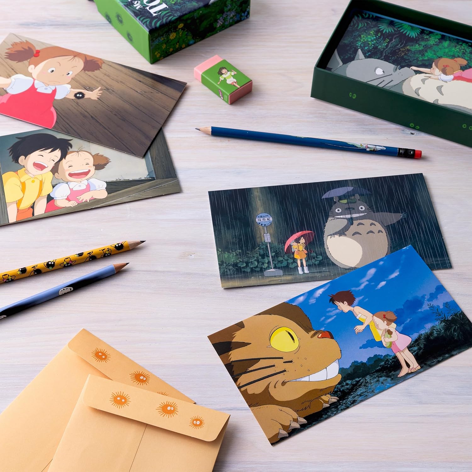 A flat lay display of a "My Neighbor Totoro" stationery set featuring illustrated notecards, tan envelopes, pencils, an eraser, and the green storage box spread across a wooden surface.