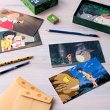 A flat lay display of a "My Neighbor Totoro" stationery set featuring illustrated notecards, tan envelopes, pencils, an eraser, and the green storage box spread across a wooden surface.