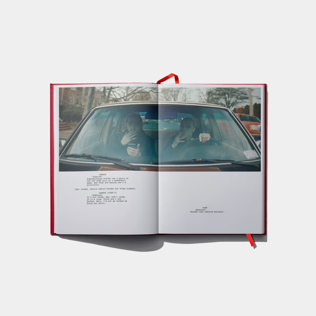 An open book displays a two-page spread featuring a cinematic film still of two men smoking in a car above formatted screenplay dialogue.