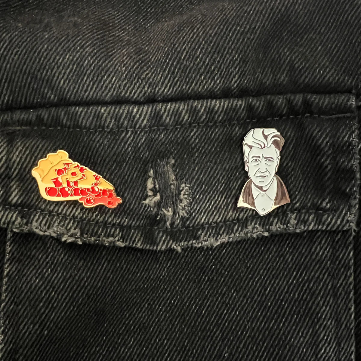 Two enamel lapel pins—one of a cherry pie slice on the left and one of David Lynch on the right—pinned to the pocket flap of a black denim jacket.