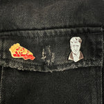 Two enamel lapel pins—one of a cherry pie slice on the left and one of David Lynch on the right—pinned to the pocket flap of a black denim jacket.
