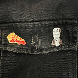 Two enamel lapel pins—one of a cherry pie slice on the left and one of David Lynch on the right—pinned to the pocket flap of a black denim jacket.