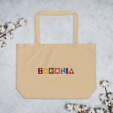 Beige coloured rectangular tote bag with top handle and printed 'Bugonia' text in multicolour at front on marbled background with dried cotton stems at each corner