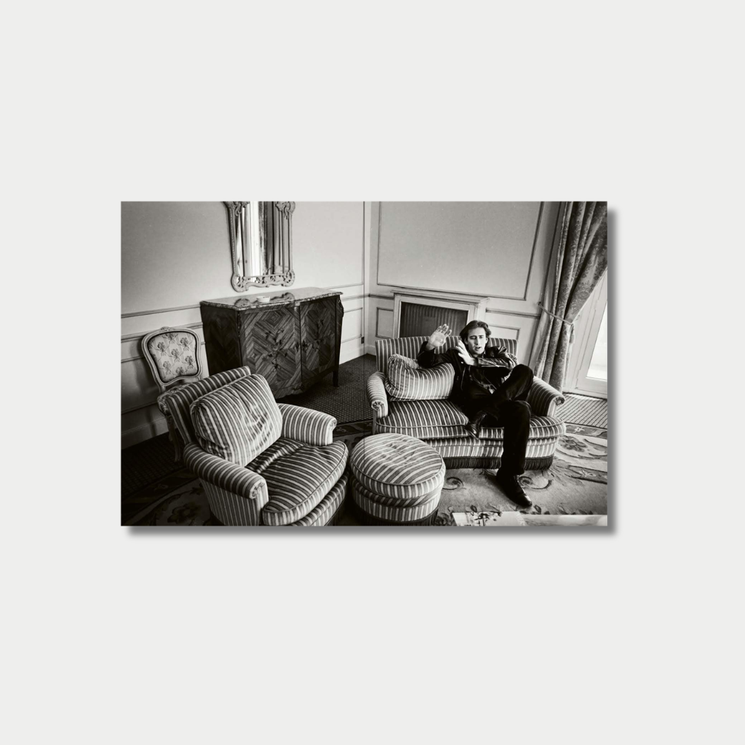 A black-and-white photograph shows actor Nicolas Cage reclining and gesturing on a striped sofa inside an elegantly decorated room featuring classic furniture, a patterned carpet, and a large window.