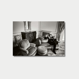 A black-and-white photograph shows actor Nicolas Cage reclining and gesturing on a striped sofa inside an elegantly decorated room featuring classic furniture, a patterned carpet, and a large window.