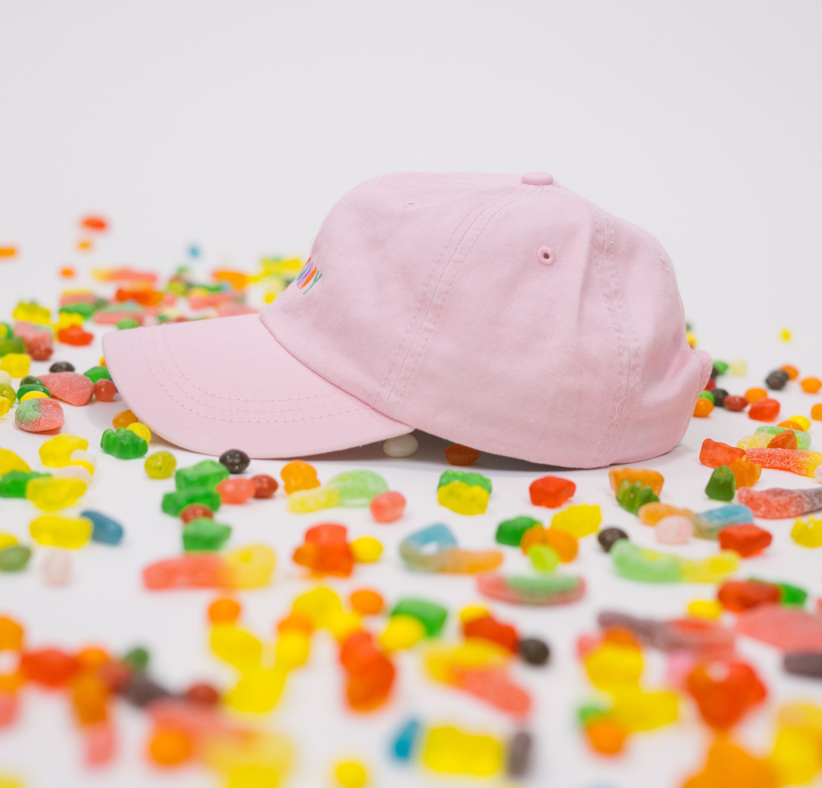 Side profile of pink baseball cap on a white background with assorted candies, featuring “Candy” embroidered in multicolour on the front.