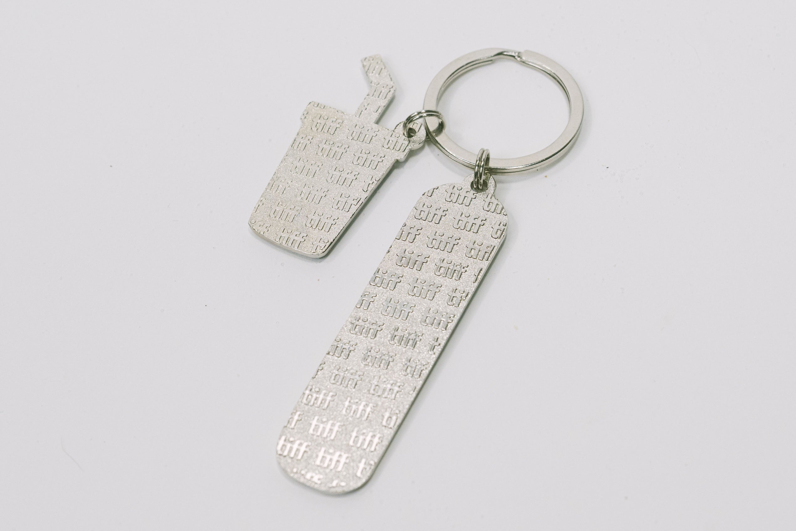Silver-tone keychain on a white background featuring back of two "TIFF" embossed charms.