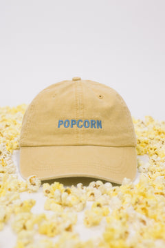 Yellow baseball cap on a white background scattered with popcorn, featuring “Popcorn” embroidered in blue on the front.