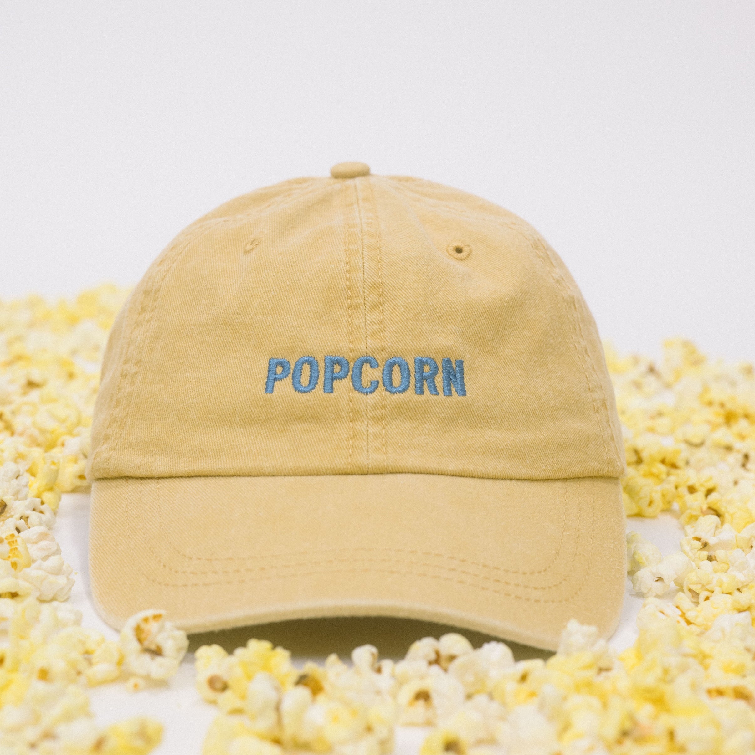 Yellow baseball cap on a white background scattered with popcorn, featuring “Popcorn” embroidered in blue on the front.