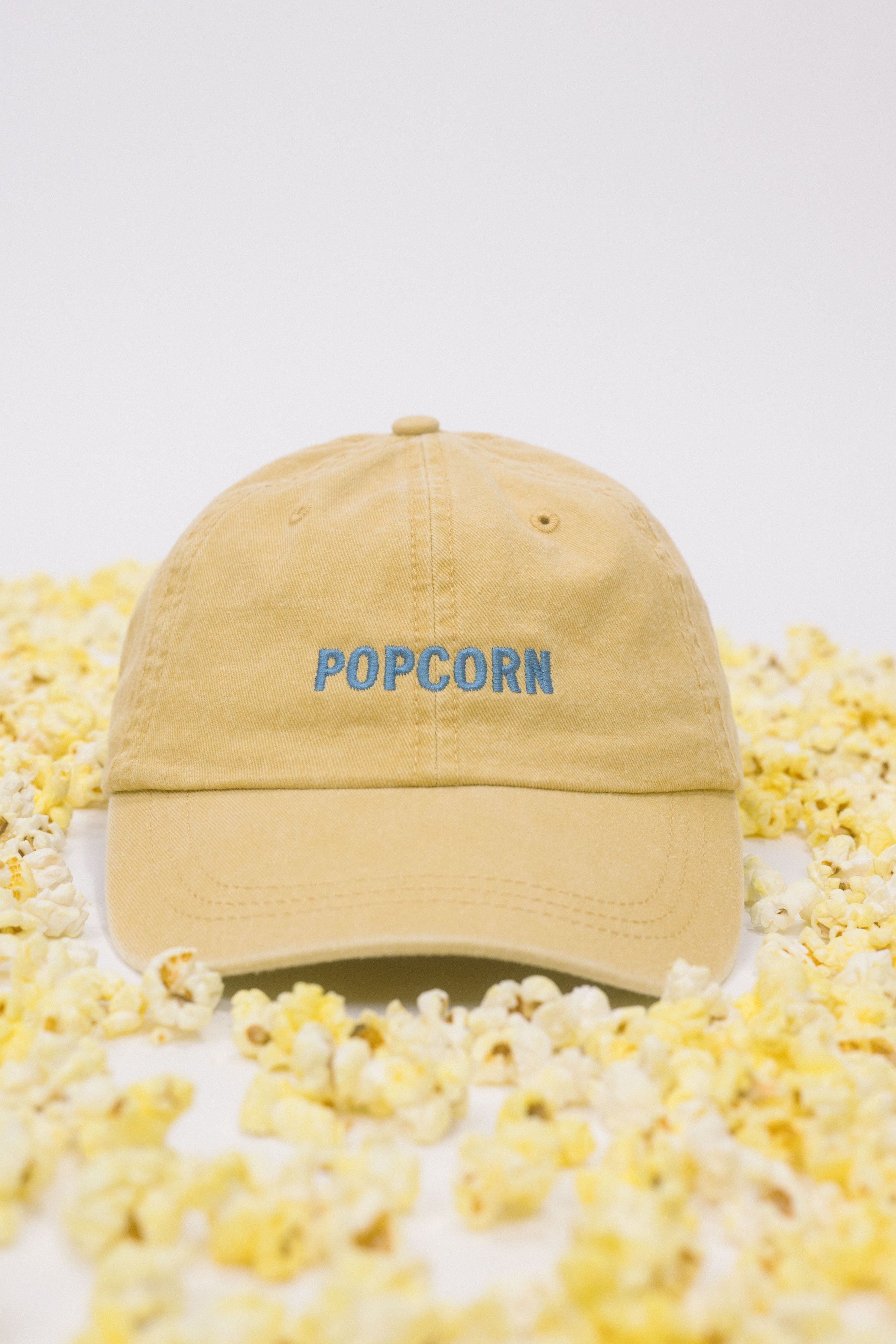 Yellow baseball cap on a white background scattered with popcorn, featuring “Popcorn” embroidered in blue on the front.