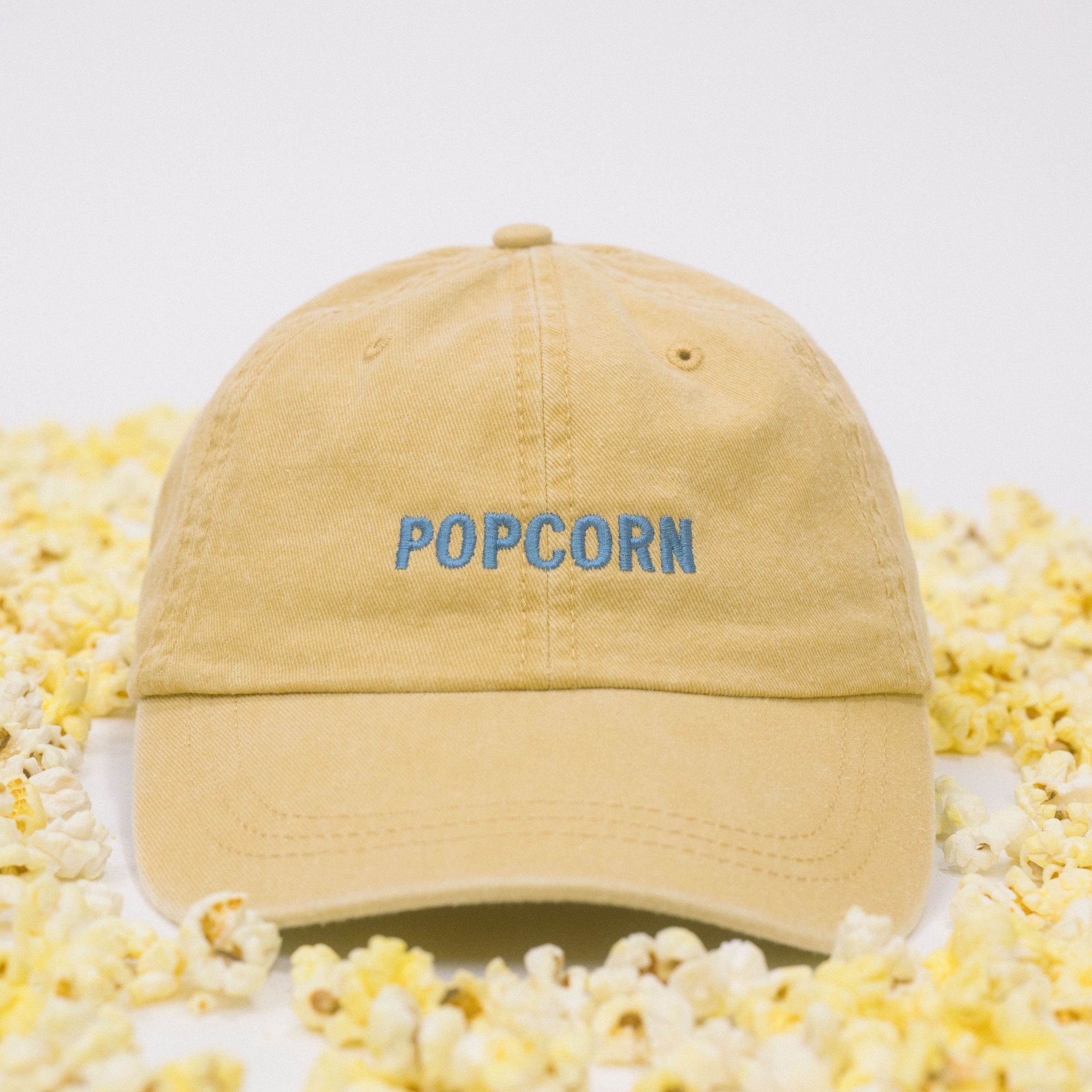 Yellow baseball cap on a white background scattered with popcorn, featuring “Popcorn” embroidered in blue on the front.