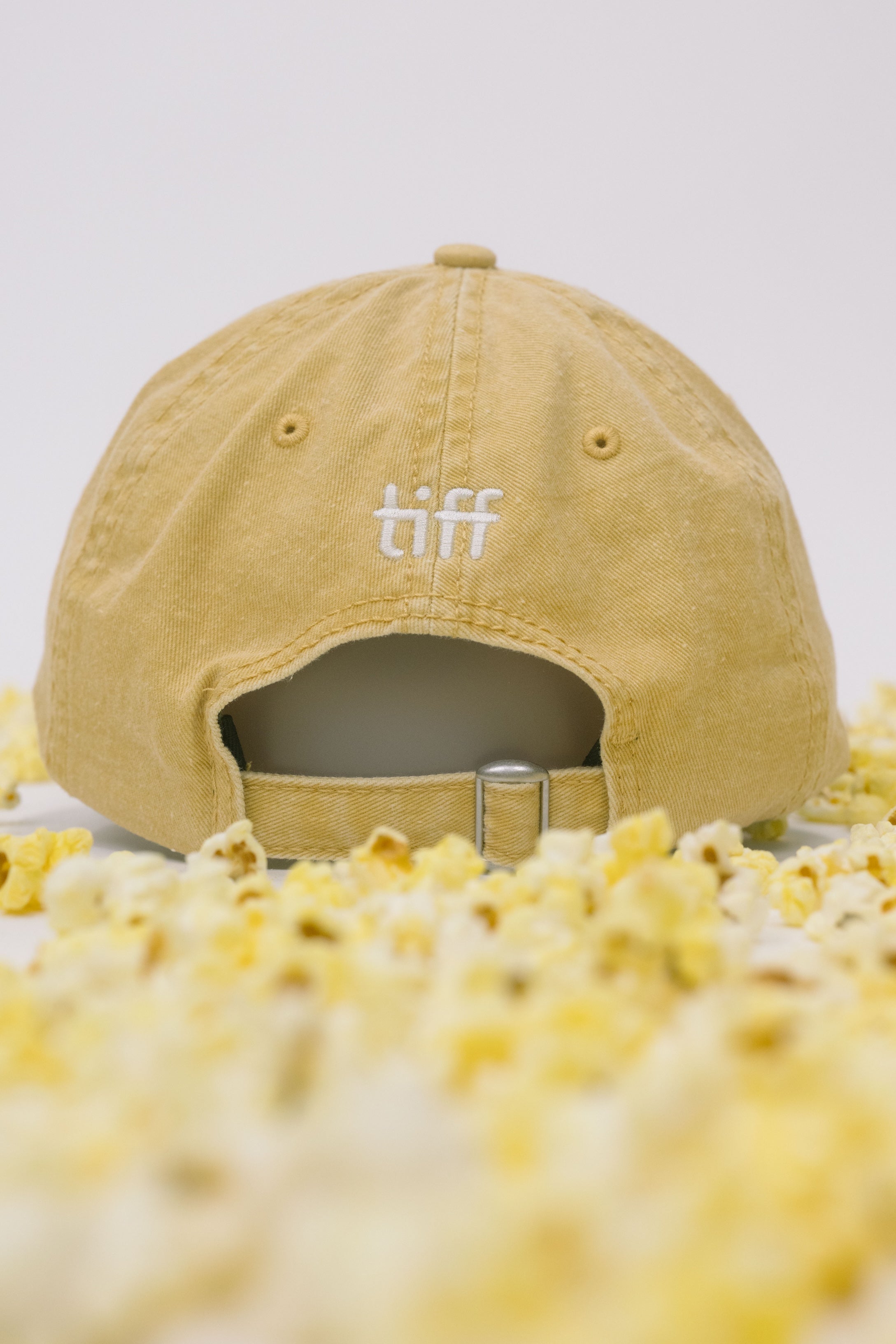 Back view of yellow baseball cap on a white background with popcorn surrounding it, featuring “TIFF” embroidery in white. 