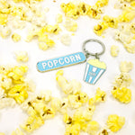 Silver-tone keychain on a white background scattered with popcorn, featuring two charms: a blue enamel “Popcorn” tag and a blue striped TIFF popcorn bucket.