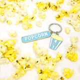 Silver-tone keychain on a white background scattered with popcorn, featuring two charms: a blue enamel “Popcorn” tag and a blue striped TIFF popcorn bucket.