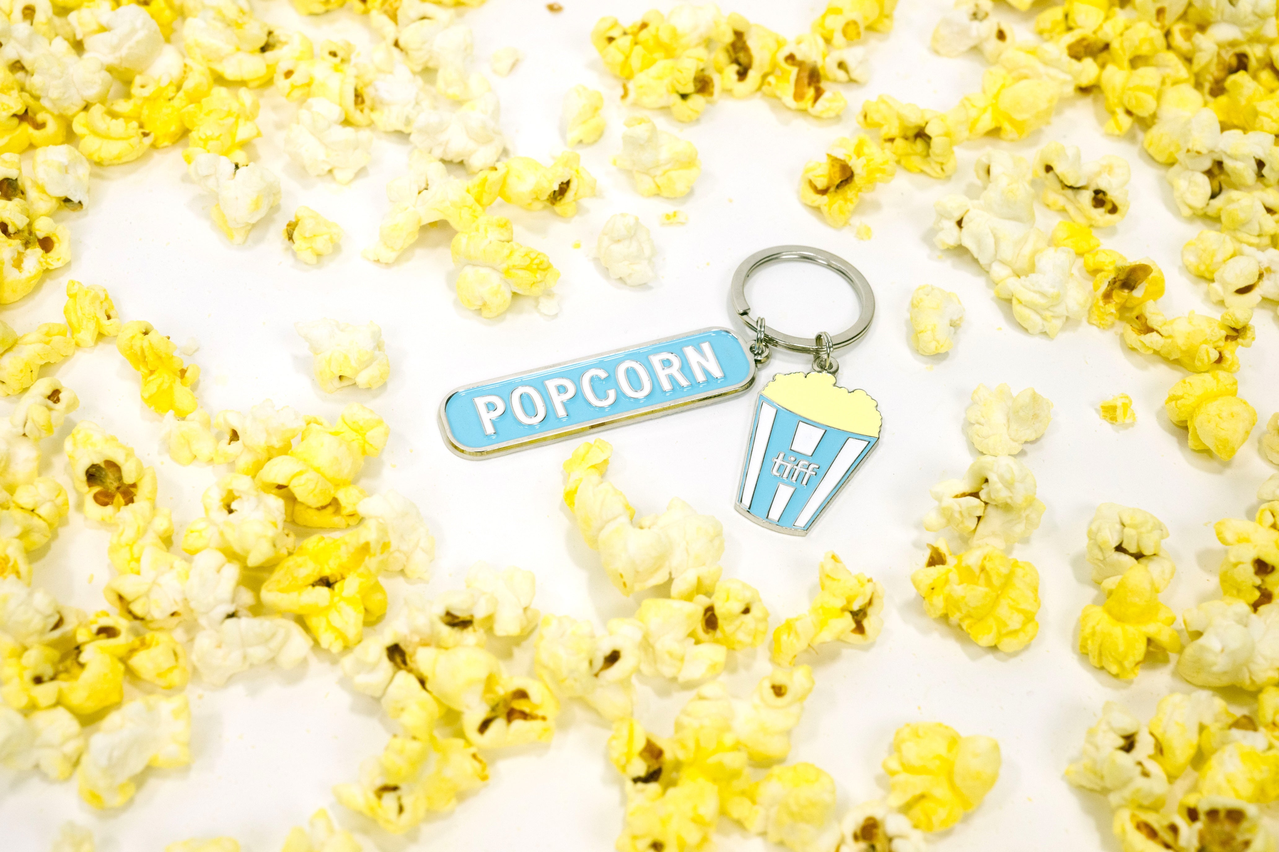 Silver-tone keychain on a white background scattered with popcorn, featuring two charms: a blue enamel “Popcorn” tag and a blue striped TIFF popcorn bucket.