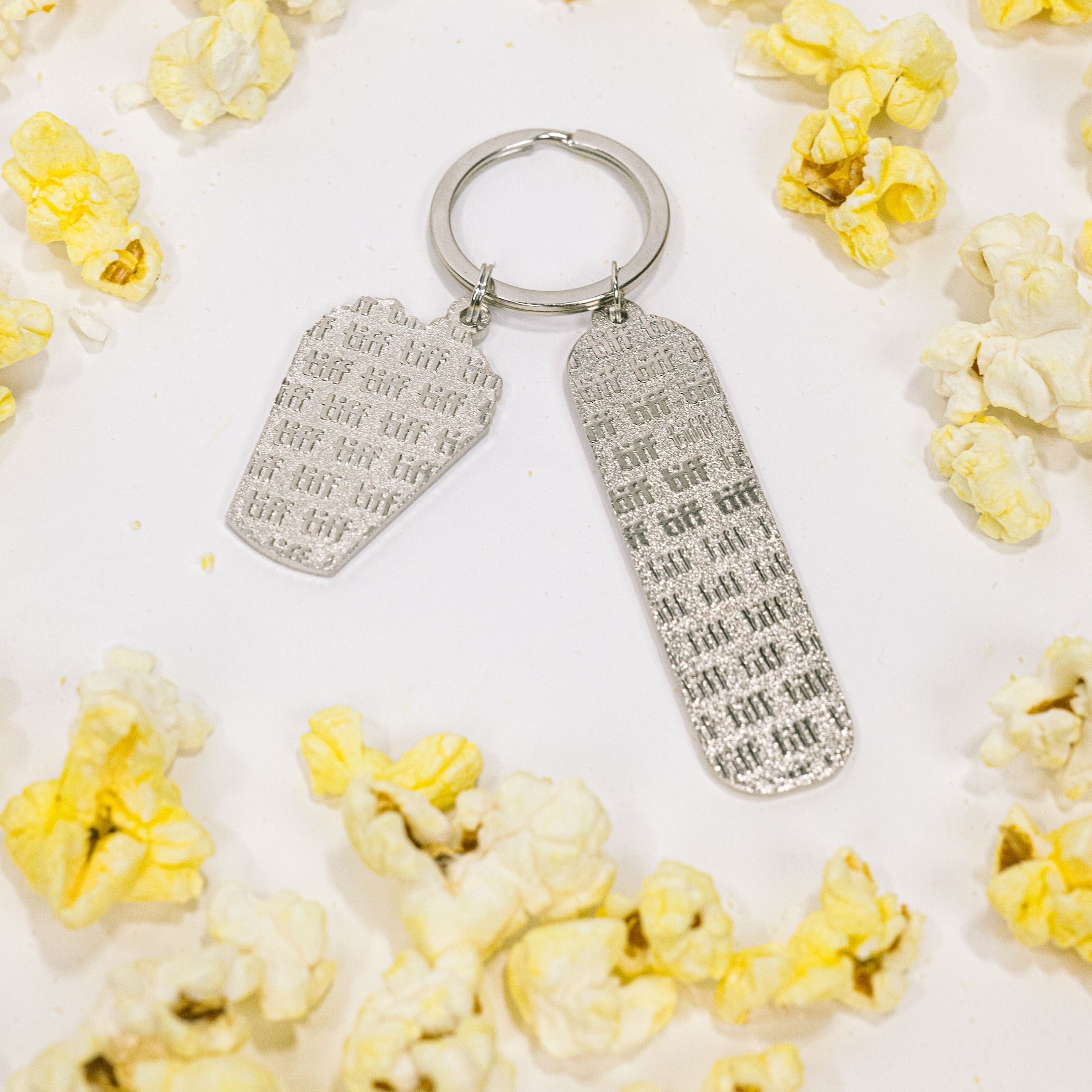 Silver-tone keychain on a white background featuring back of two "TIFF" embossed charms surrounded by yellow popcorn.