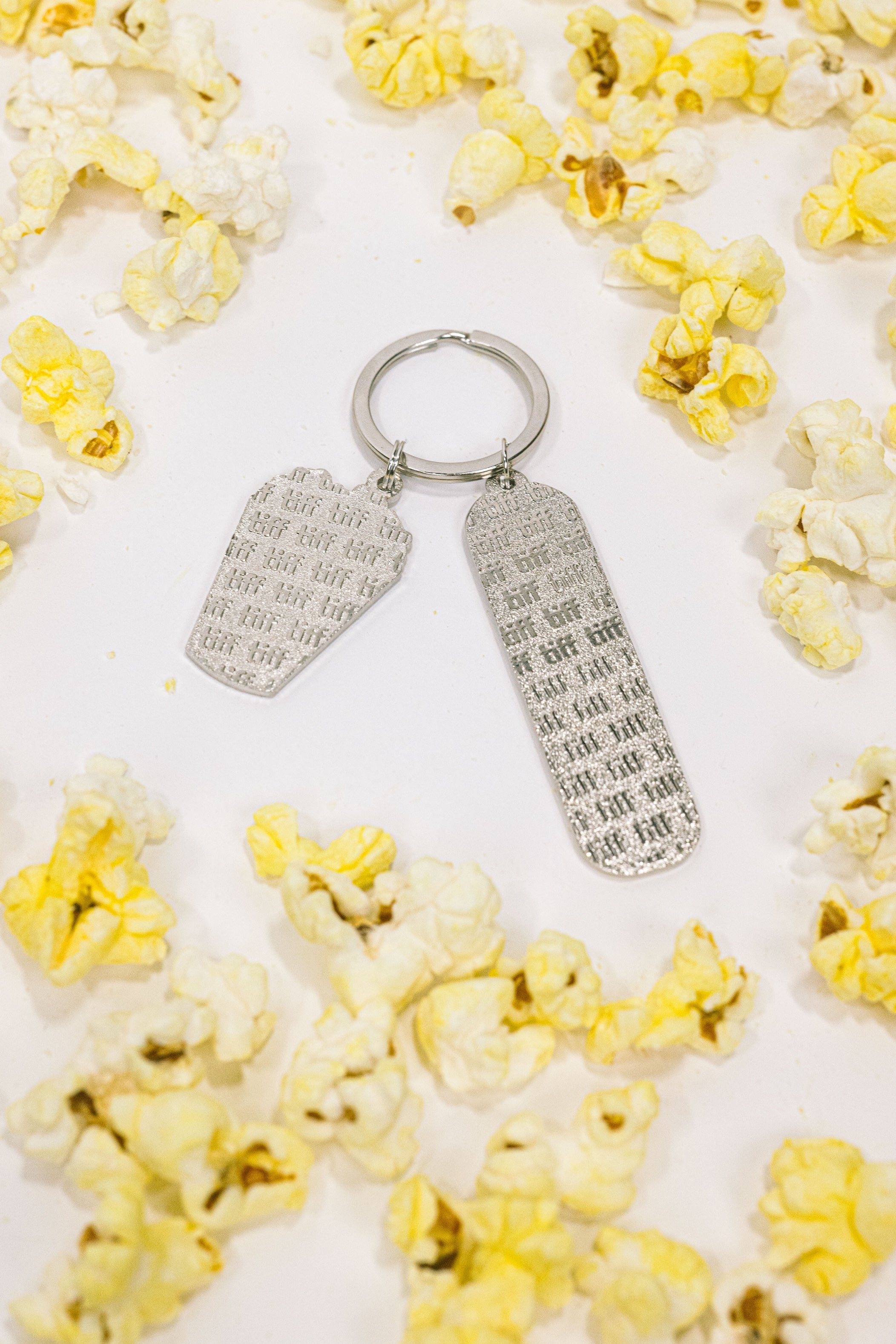 Silver-tone keychain on a white background featuring back of two "TIFF" embossed charms surrounded by yellow popcorn.