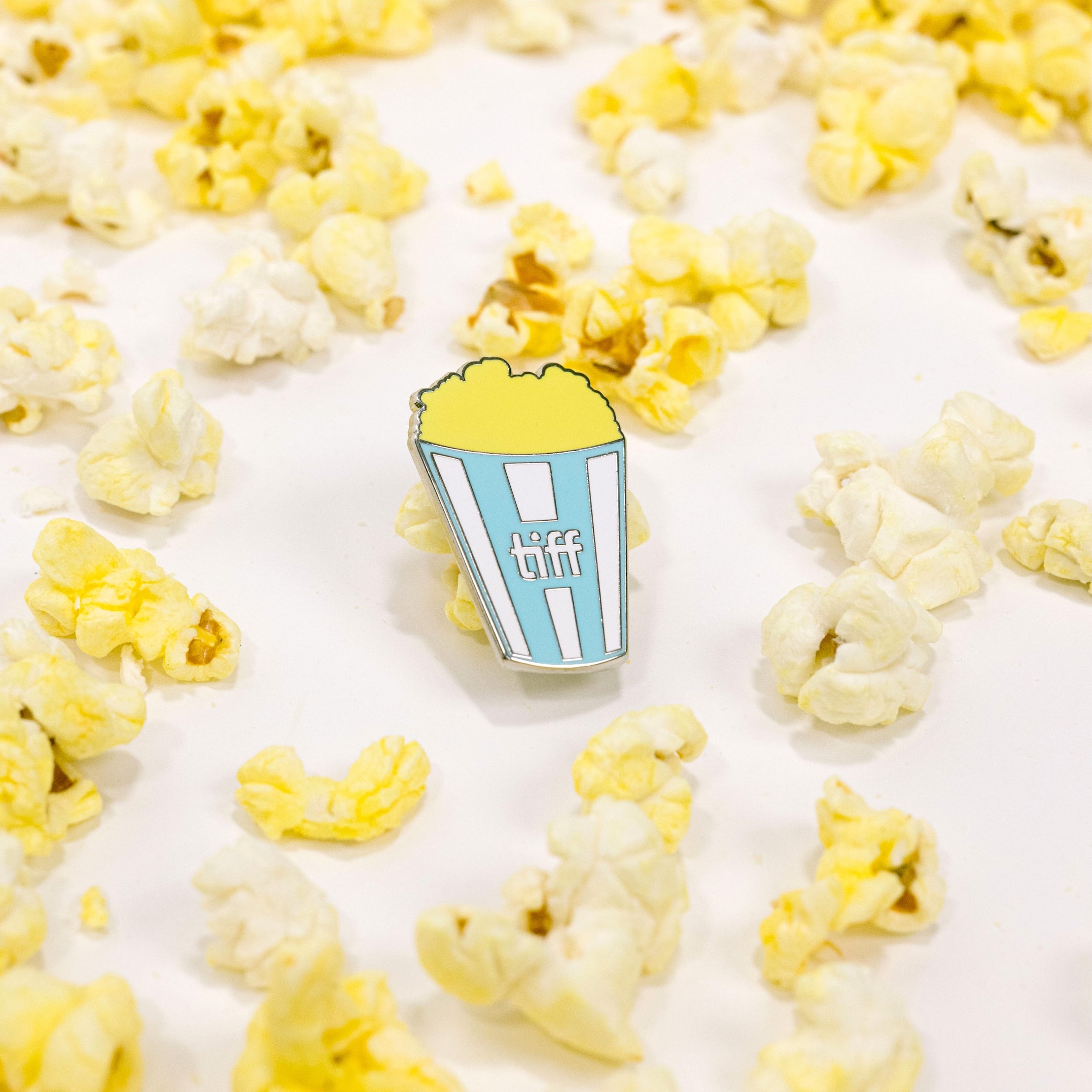 Pin with a blue and white striped popcorn container surrounded by yellow popcorn on a white background