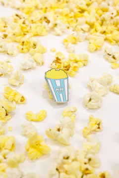 Pin with a blue and white striped popcorn container surrounded by yellow popcorn on a white background