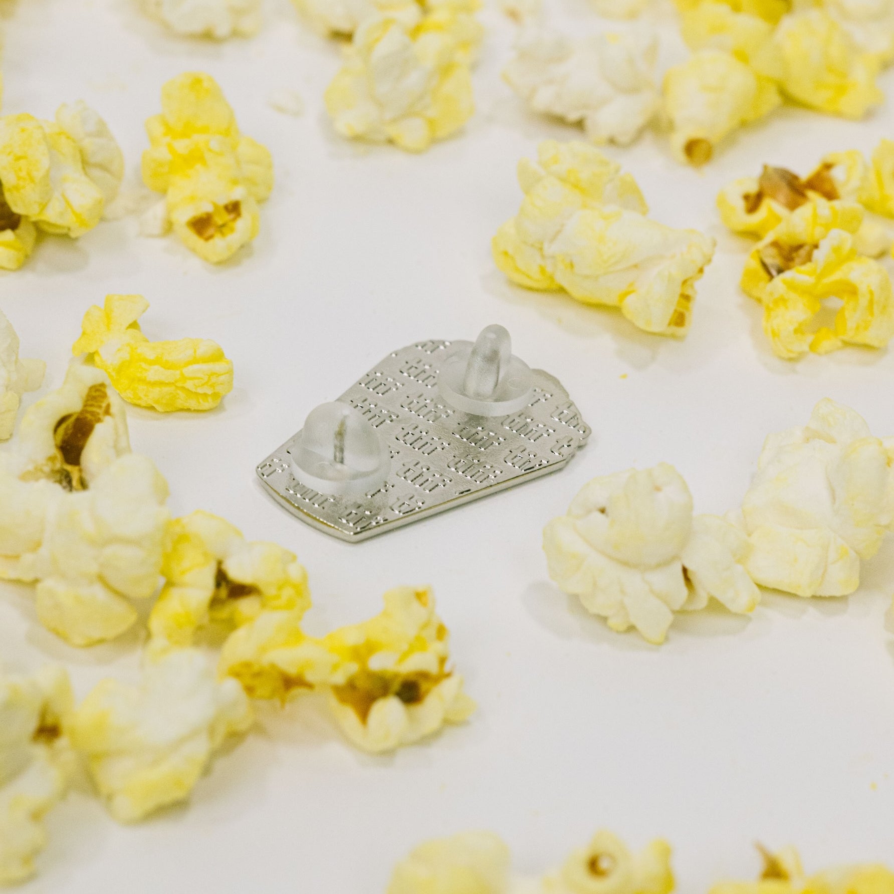 Back view of a silver enamel pin in silver, featuring an embossed TIFF monogram with silicone backings on a white background surrounded by yellow popcorn.