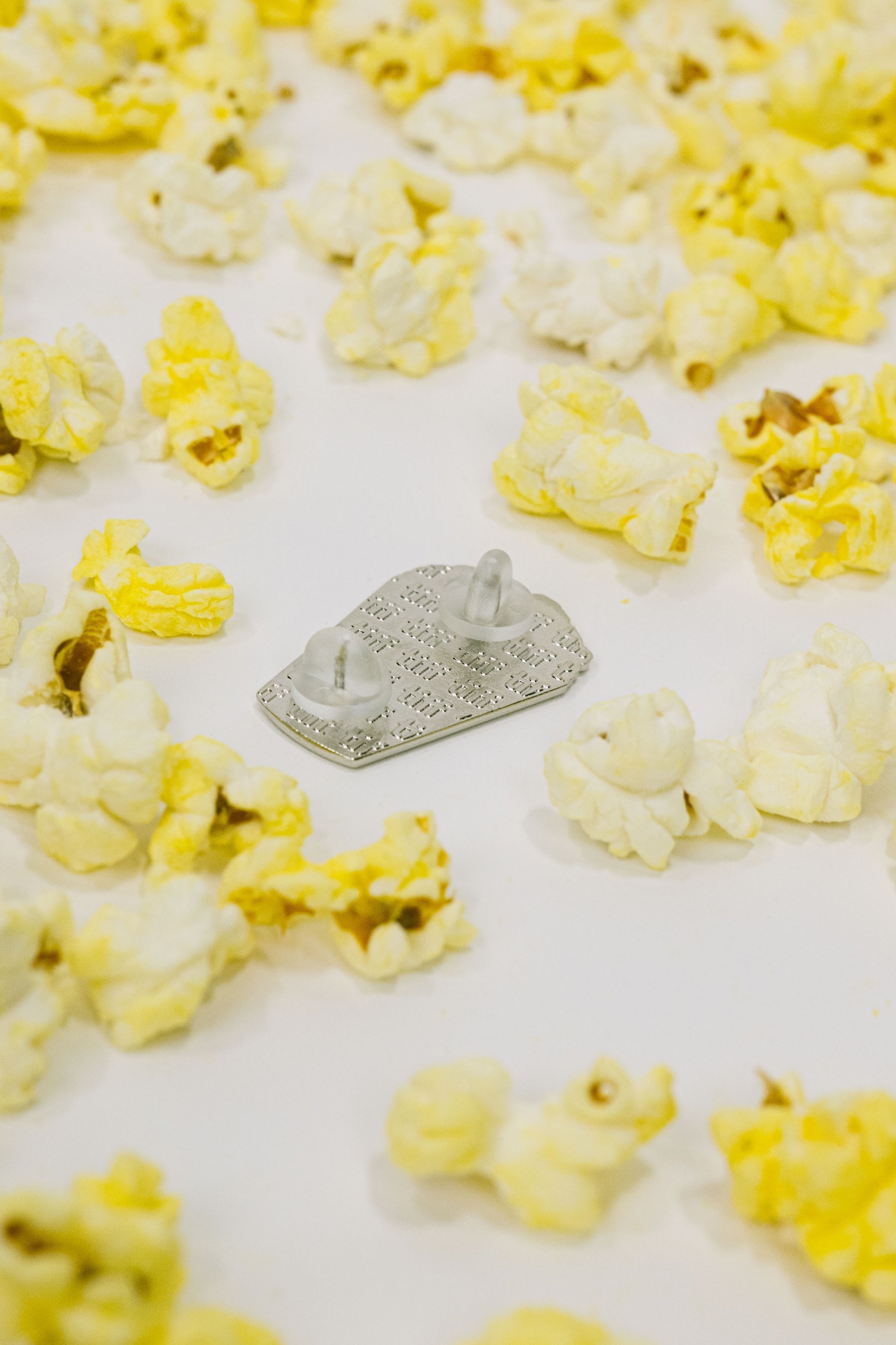 Back view of a silver enamel pin in silver, featuring an embossed TIFF monogram with silicone backings on a white background surrounded by yellow popcorn.