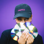 Person wearing a 'Cinephile' cap holding CDs in front of their face on a purple background