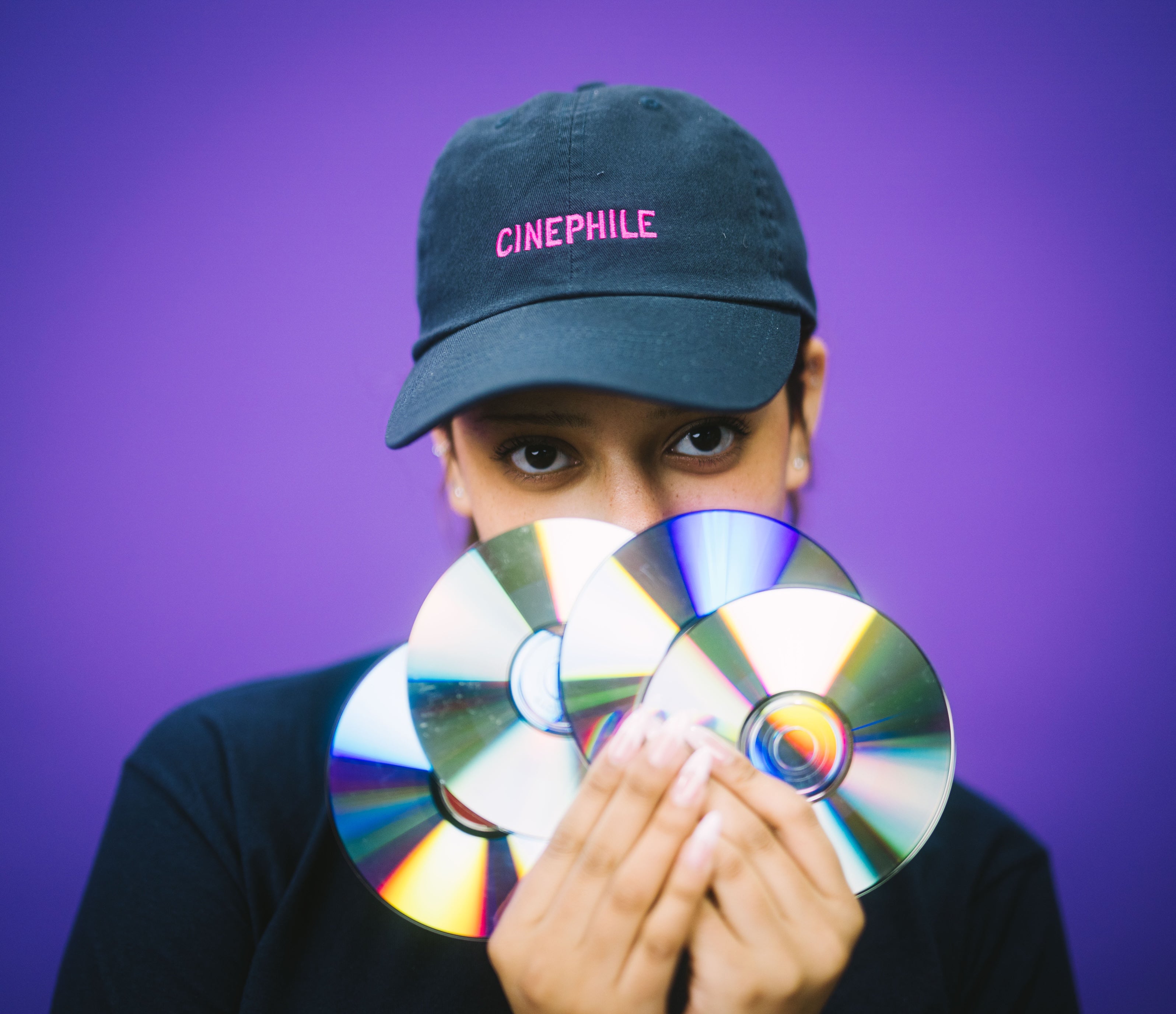 Person wearing a 'Cinephile' cap holding CDs in front of their face on a purple background