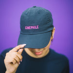 Person wearing a dark blue cap with 'CINEPHILE' text and a black t-shirt with 'tiff' text, against a purple background.