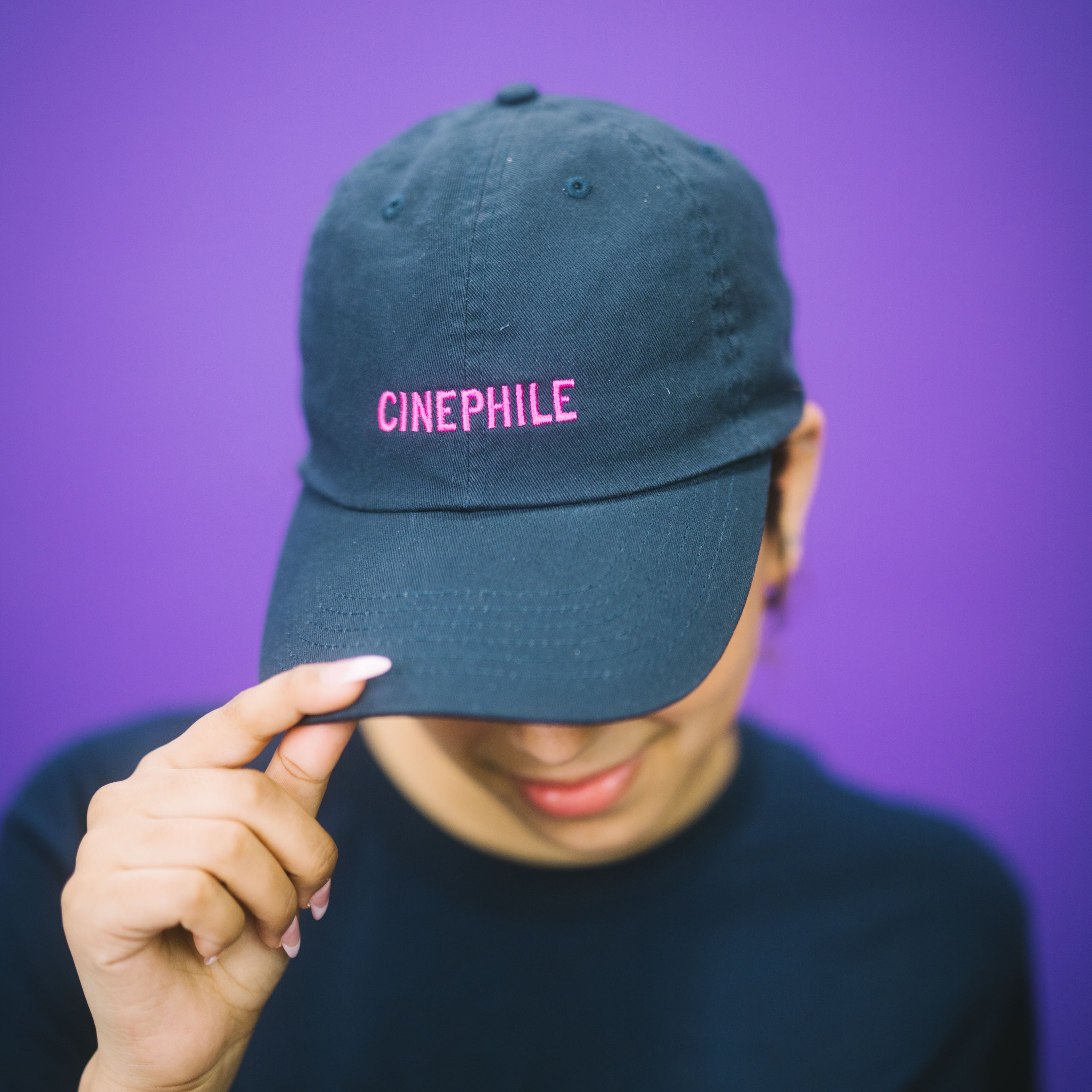 Person wearing a dark blue cap with 'CINEPHILE' text and a black t-shirt with 'tiff' text, against a purple background.