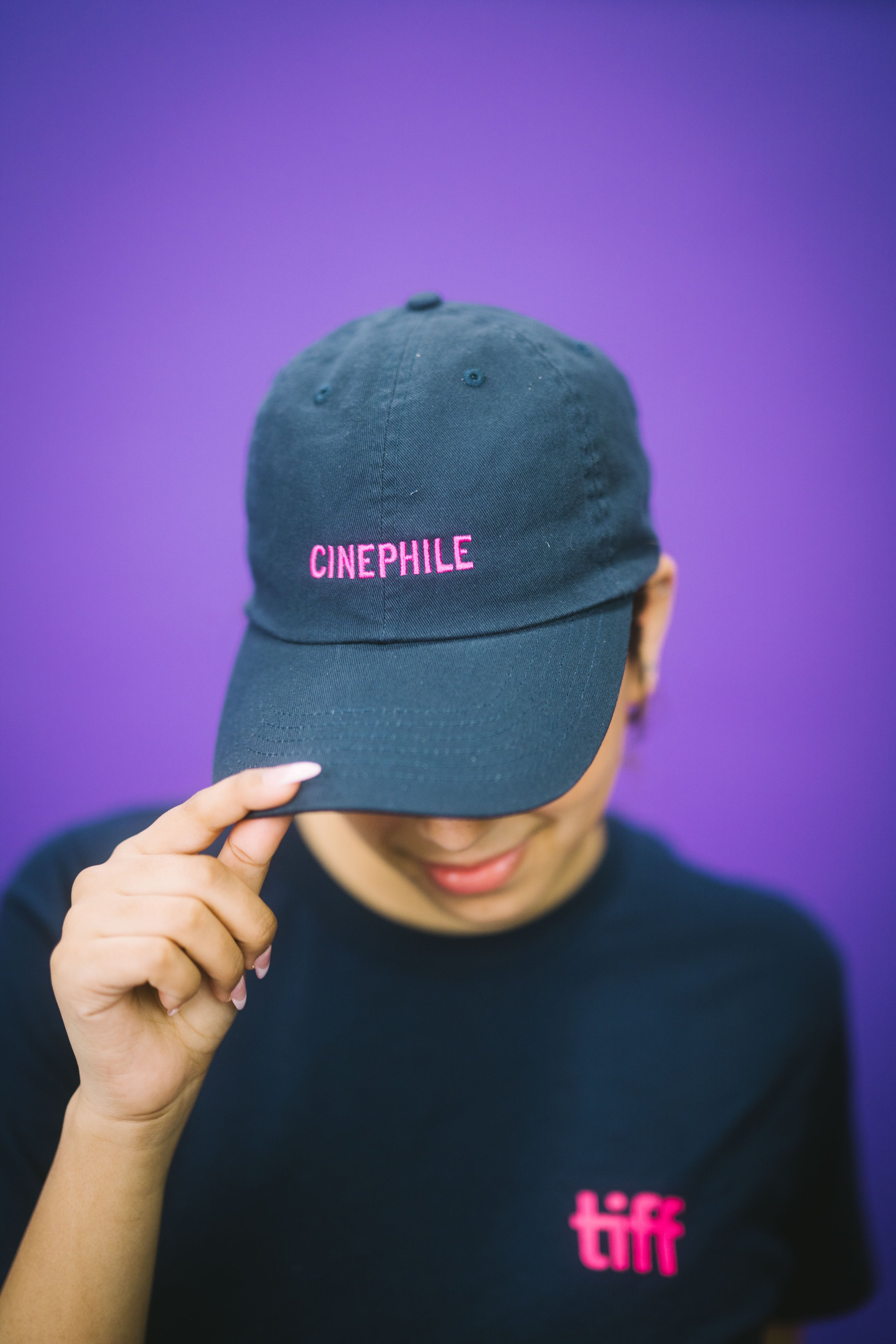 Person wearing a dark blue cap with 'CINEPHILE' text and a black t-shirt with 'tiff' text, against a purple background.
