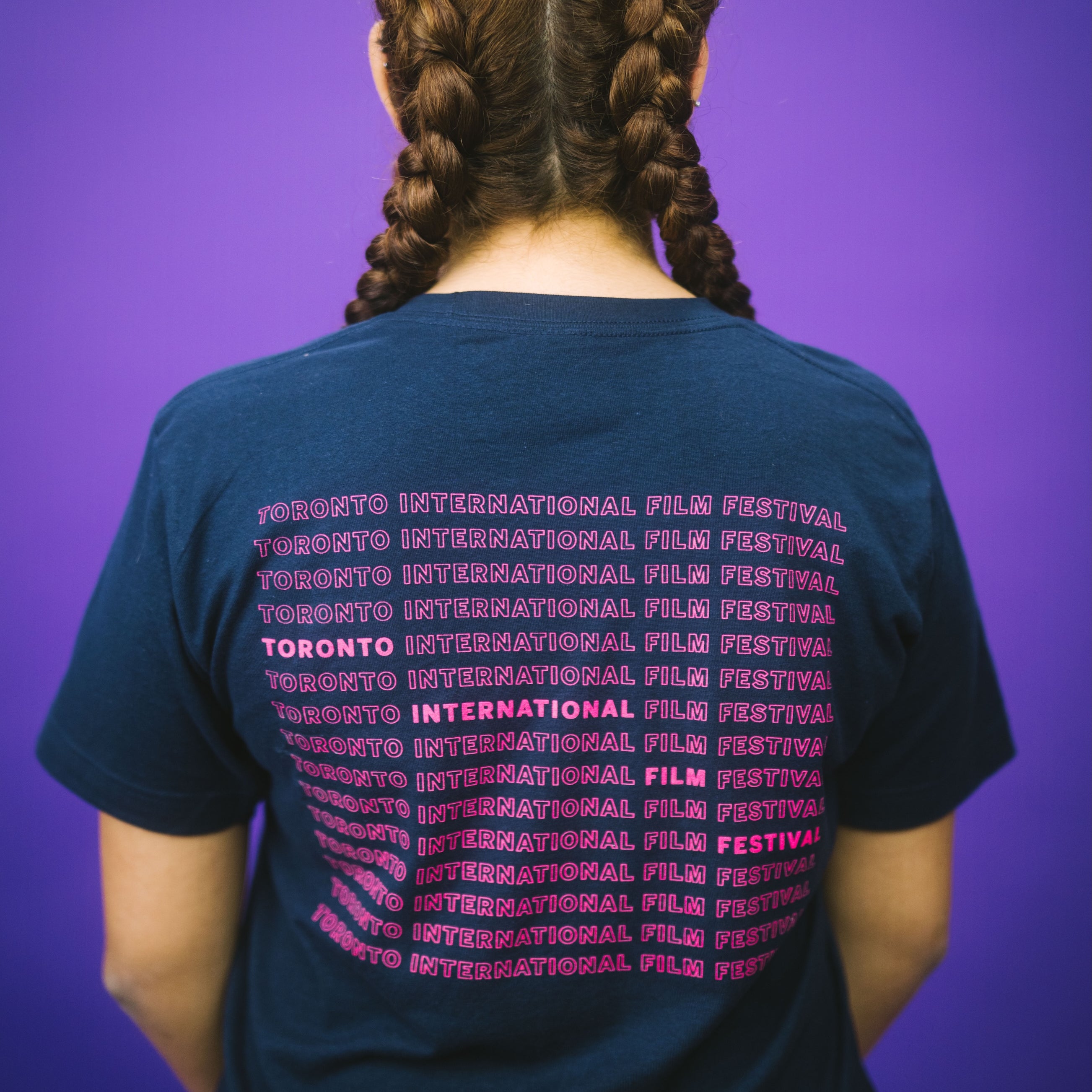 A person with brown hair dutch braids, seen from behind, wears a navy blue T-shirt featuring "Toronto International Film Festival" repeated in pink text.

