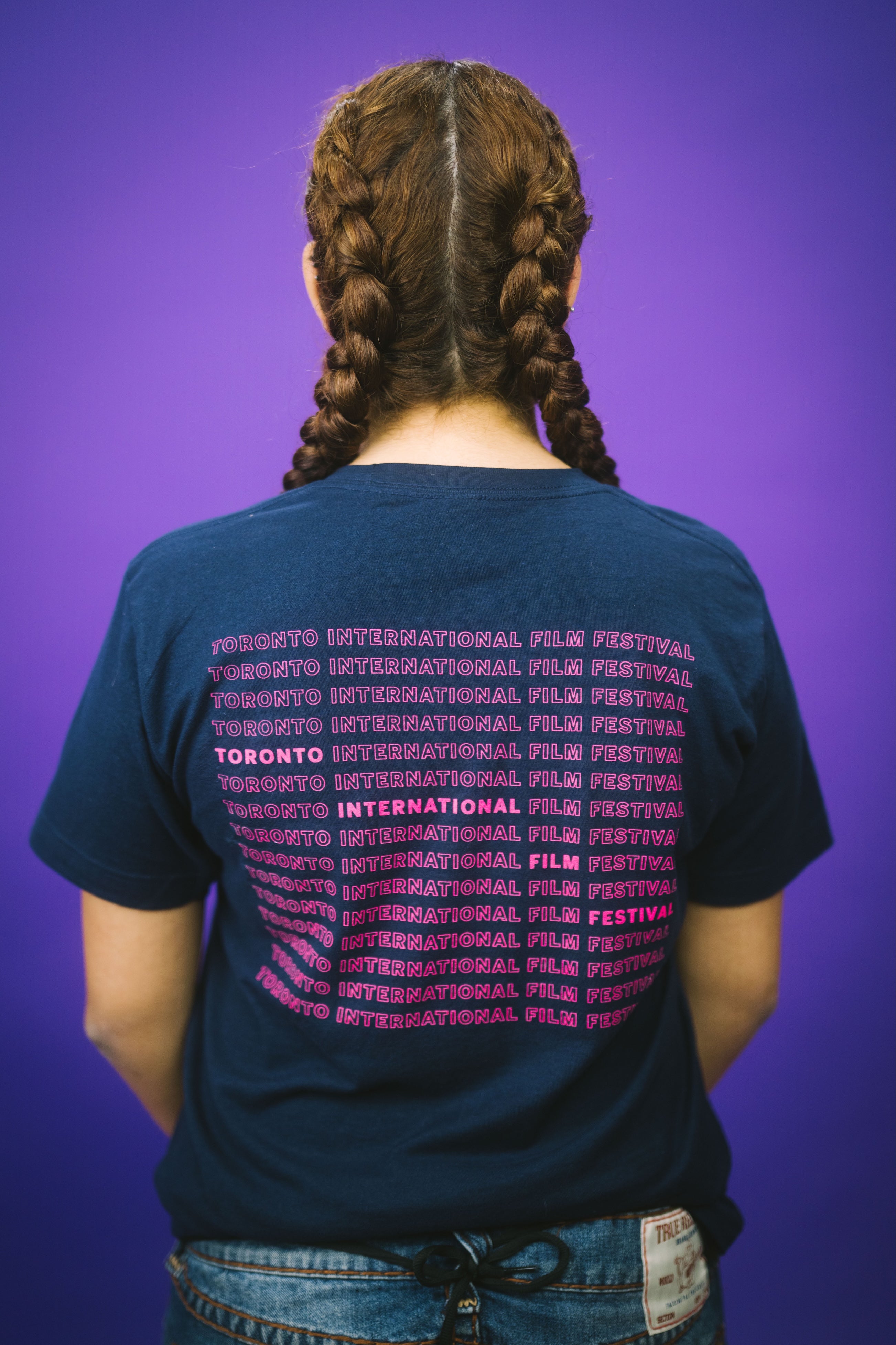 A person with brown hair dutch braids, seen from behind, wears a navy blue T-shirt featuring "Toronto International Film Festival" repeated in pink text.
