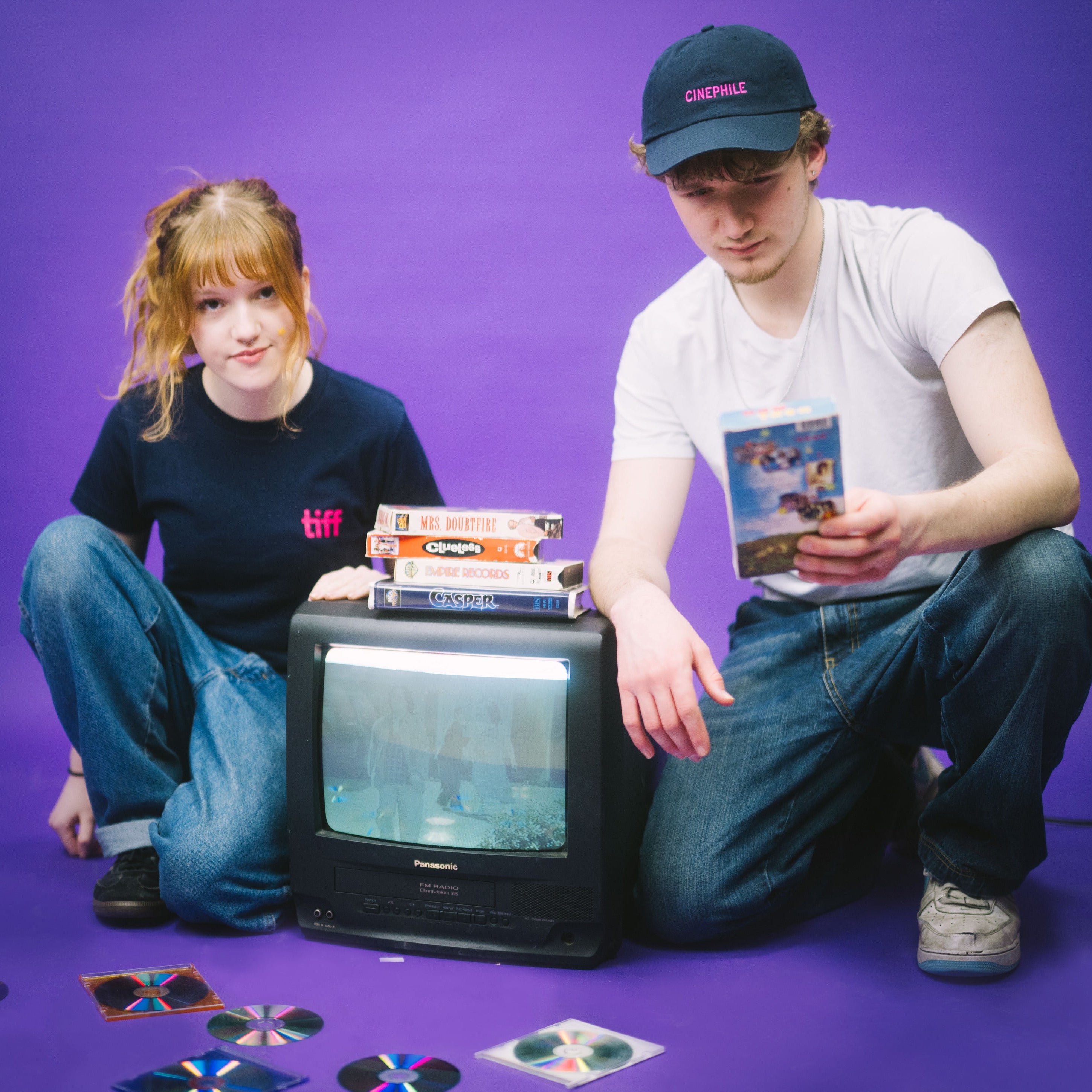 Two young people against a purple background with a VCR TV and different VHS movies stacked around them.