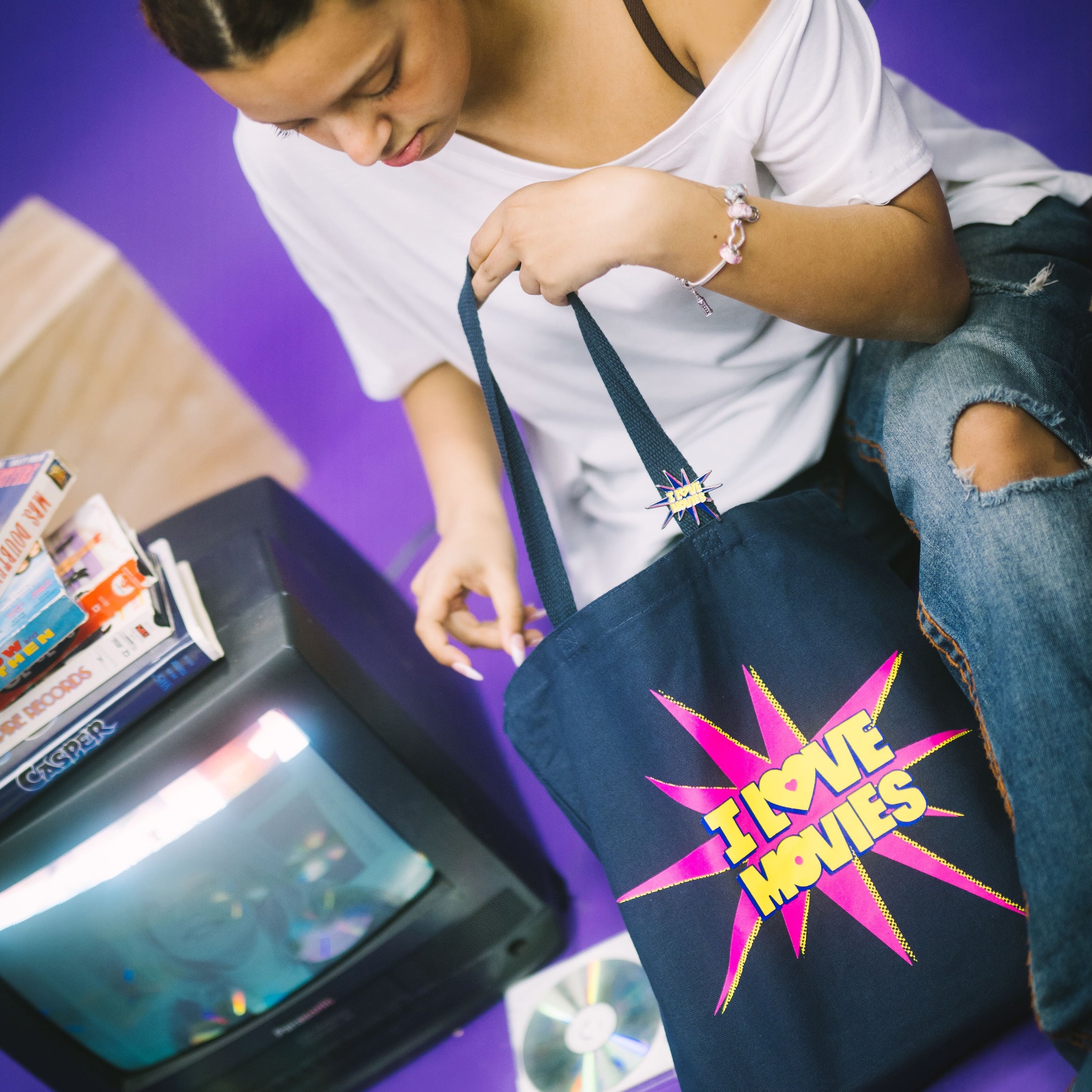 Person holding a tote bag with 'I love movies' design on a purple floor with a vintage television and CDs.