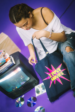 Person holding a tote bag with 'I love movies' design on a purple floor with a vintage television and CDs.