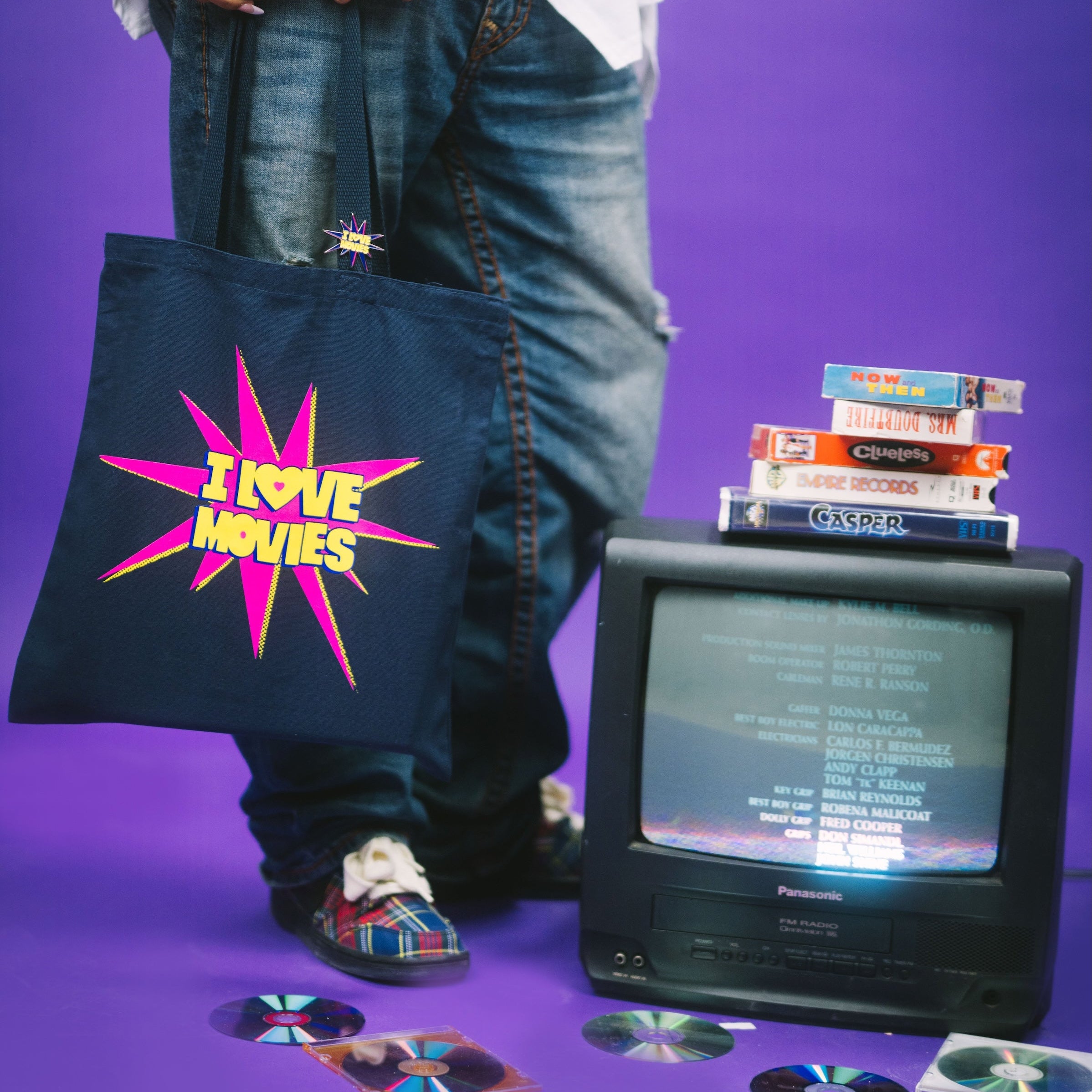 Person holding a tote bag with 'I love movies' design, standing next to a small television and CDs on a purple background.
