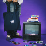 Person holding a tote bag with 'tiff' logo, vintage TV with stacked books, and scattered CDs on a purple background