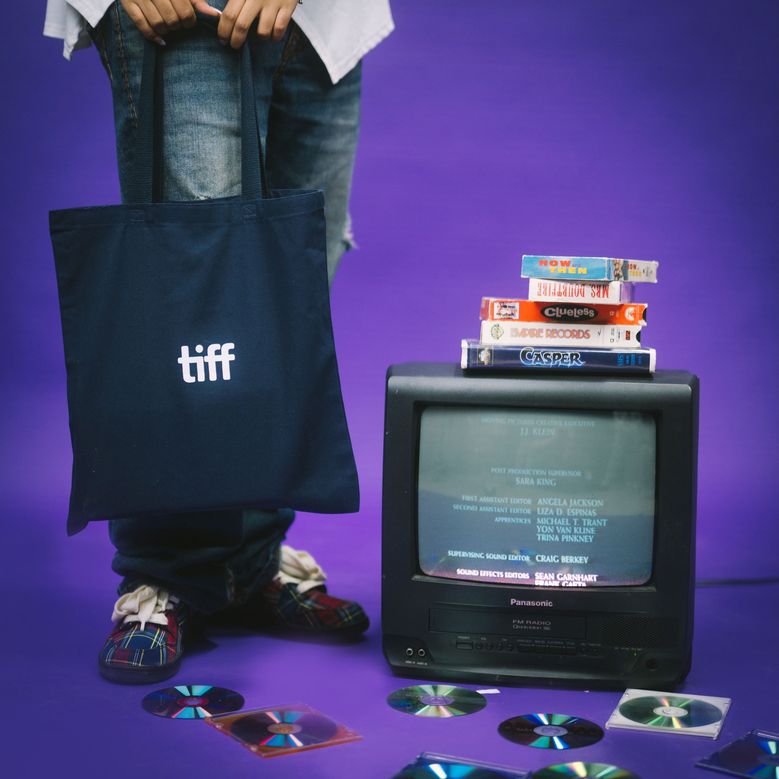 Person holding a tote bag with 'tiff' logo, vintage TV with stacked books, and scattered CDs on a purple background