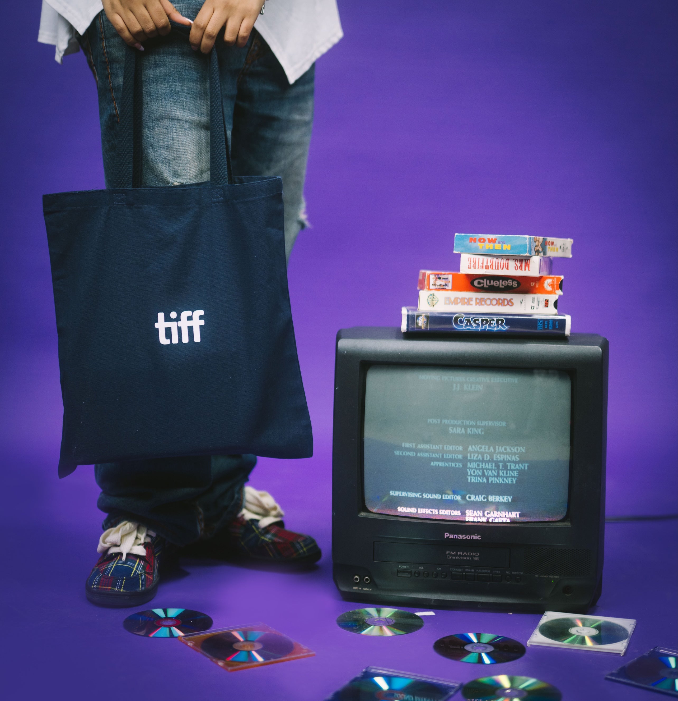 Person holding a tote bag with 'tiff' logo, vintage TV with stacked books, and scattered CDs on a purple background