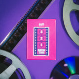 Pink 'tiff' Next Wave magnet with film reels on a purple background