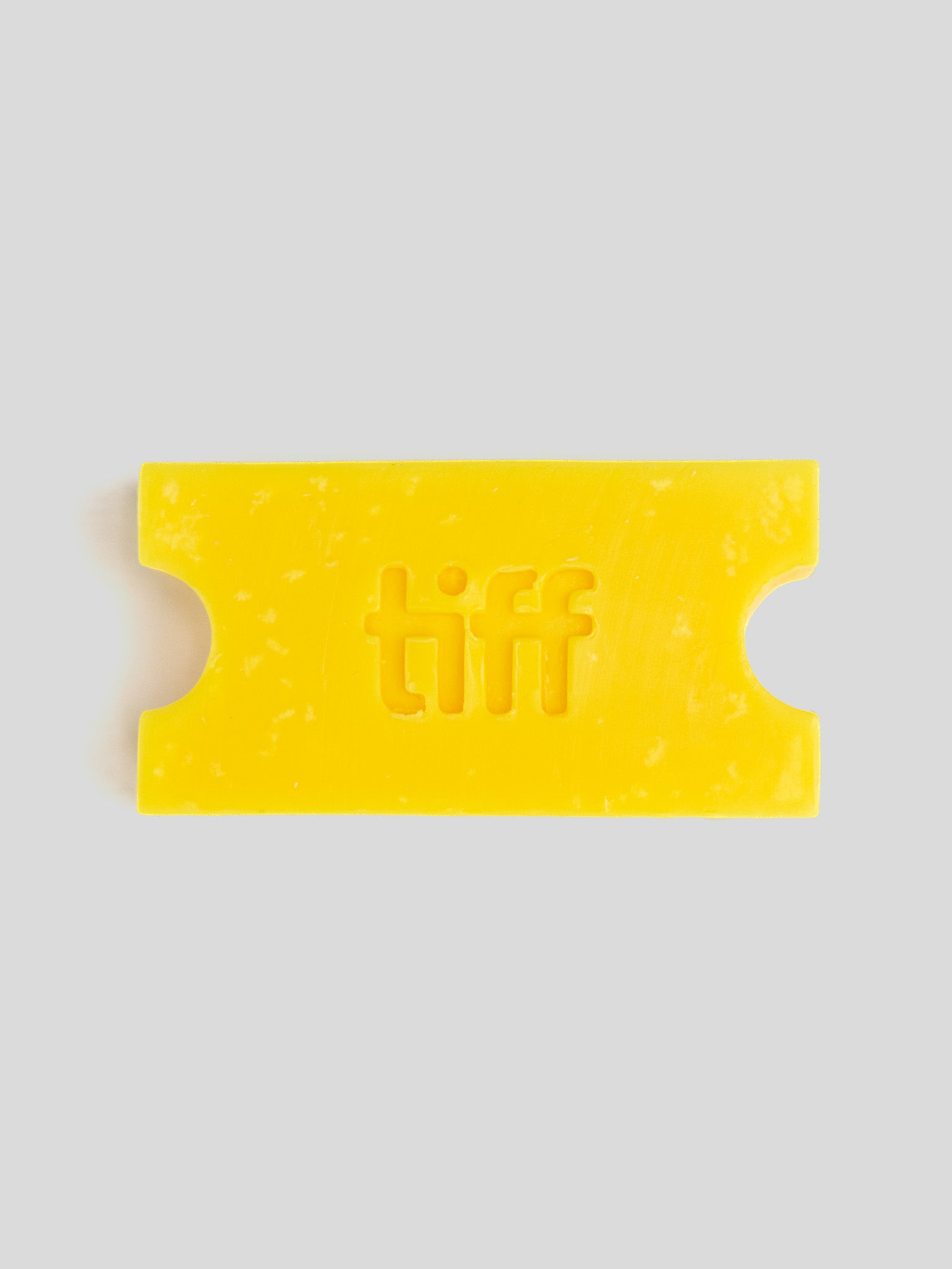 A bright yellow wax melt or soap bar shaped like a movie ticket, embossed with the word 'tiff' in the center, on a solid light gray background.