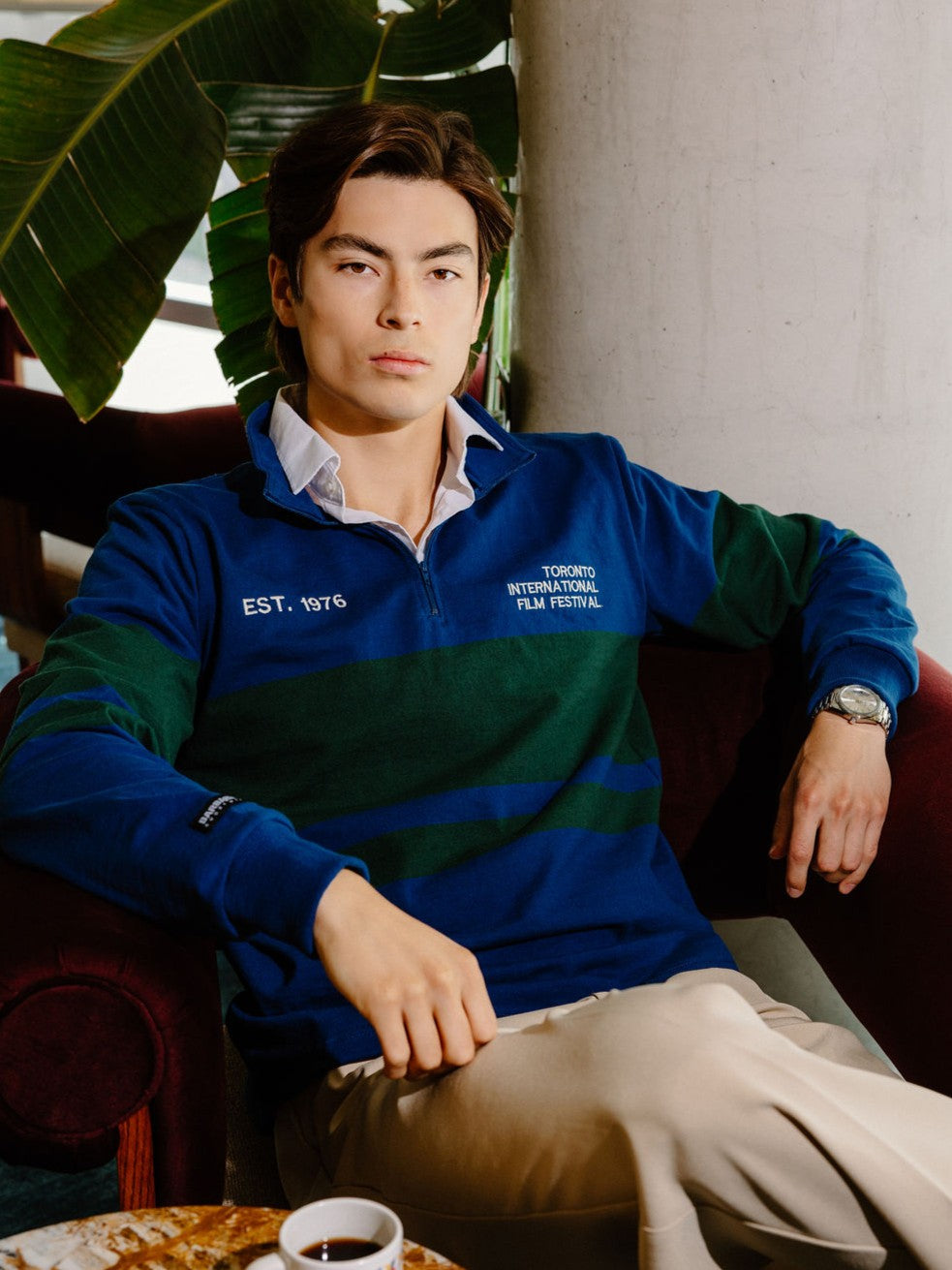 A man wearing a blue and green striped quarter-zip sweatshirt with white embroidered text, sitting in a dark red armchair in a cafe-like setting.