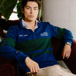 A man wearing a blue and green striped quarter-zip sweatshirt with white embroidered text, sitting in a dark red armchair in a cafe-like setting.